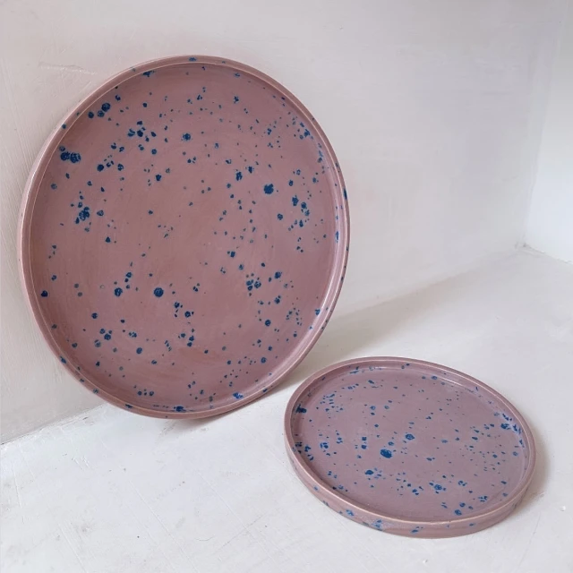 Two pink ceramic plates with blue speckles sit on a white surface, one large and one smaller, creating a minimalist, decorative scene.