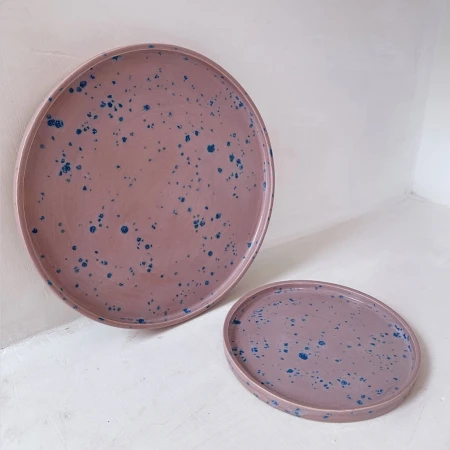 Two pink ceramic plates with blue speckles sit on a white surface, one large and one smaller, creating a minimalist, decorative scene.