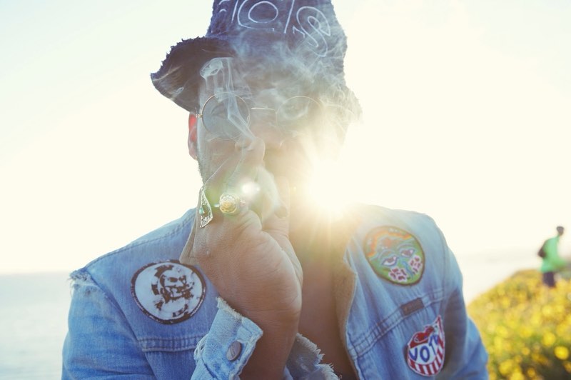 A person in a bucket hat and denim jacket, partially obscured by smoke, standing outdoors near an ocean with yellow flowers.