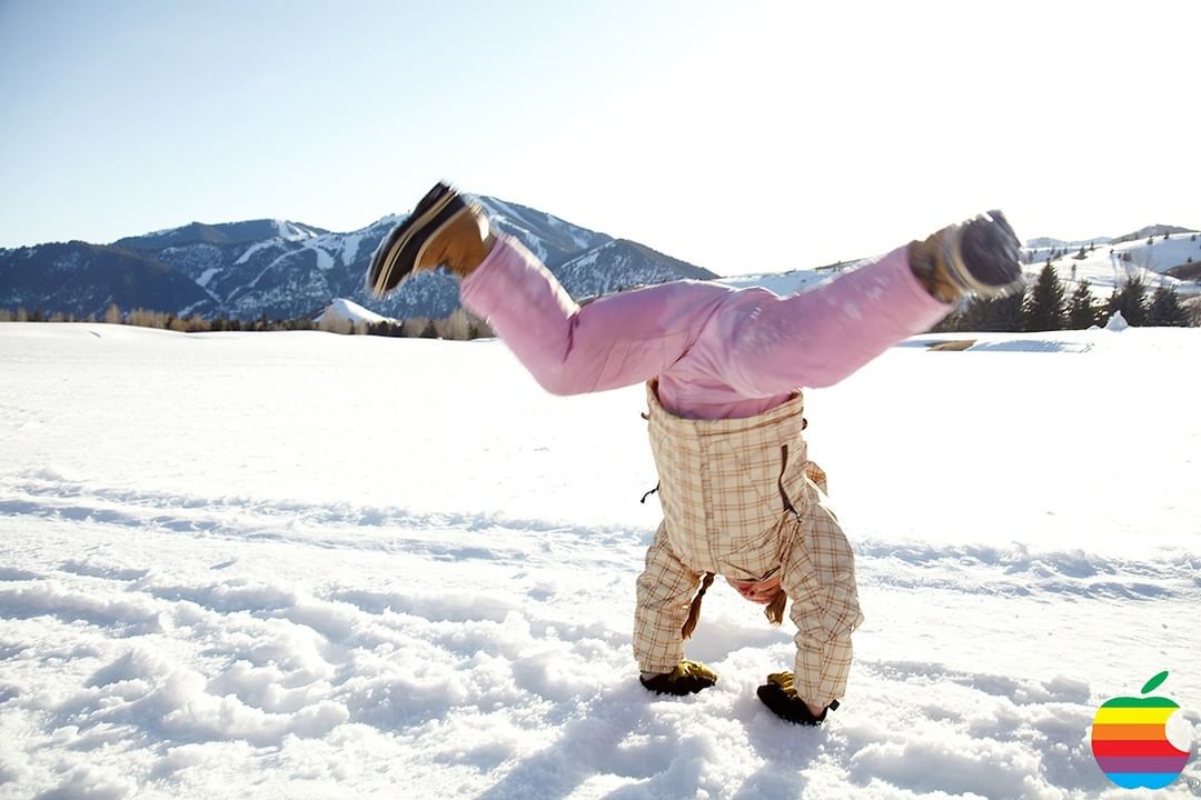 A child in pink snow pants and winter boots does a handstand in a snowy field on a bright, sunny day, with mountains in background.