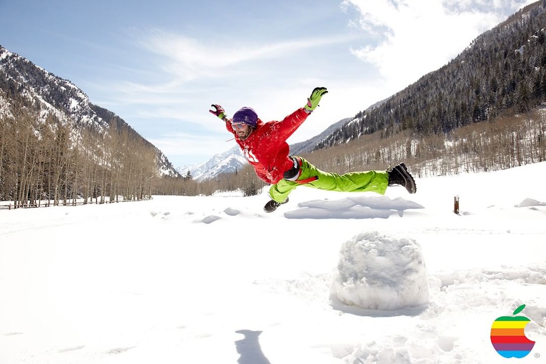 Action shot of an adult male in a red jacket and green pants doing a snow angel in front of a snowy mountain landscape on a sunny day.