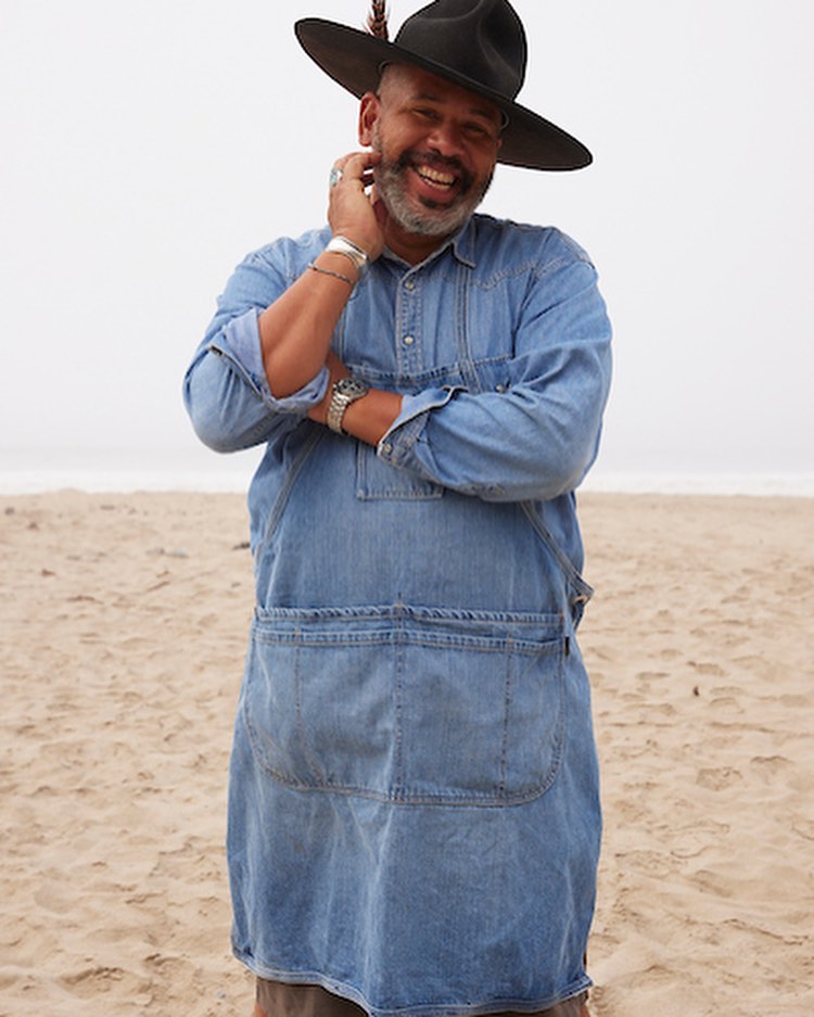 Man in denim apron and black hat smiles on beach with ocean backdrop. Casual fashion and outdoor setting. Natural light, candid portrait.