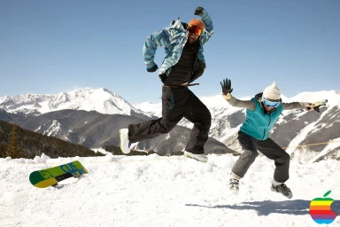 Two people jumping mid-air in a snowy mountain landscape with a snowboard lying on the snow