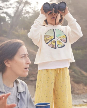 A child in a white hoodie looking through binoculars stands beside a seated woman against an outdoor hazy background.