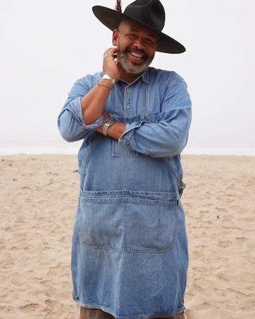 Smiling man wearing a wide-brimmed black hat and a blue denim apron on a sandy beach