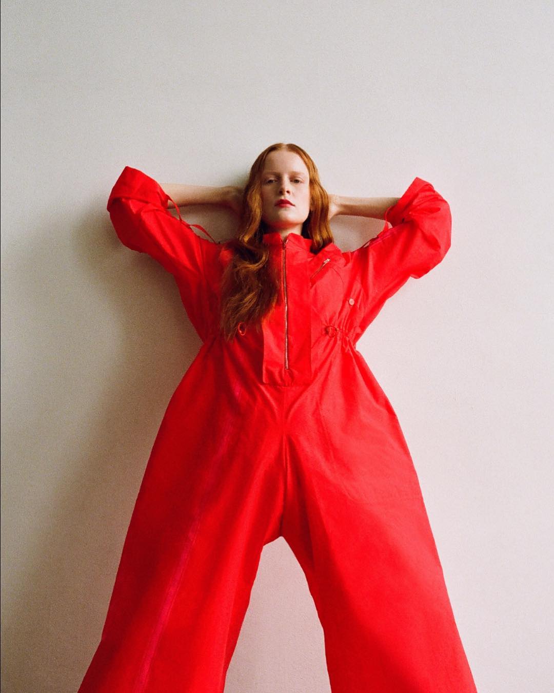 Woman with red hair in a red jumpsuit poses in front of a white wall with her hands behind her head in a studio shot.