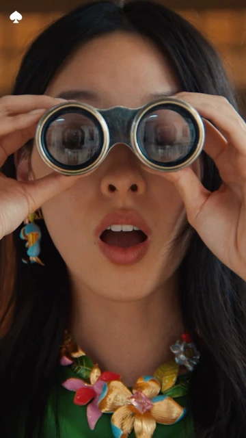 A young Asian woman gazes through binoculars with a surprised expression, wearing a vibrant floral necklace and earrings in an indoor setting.