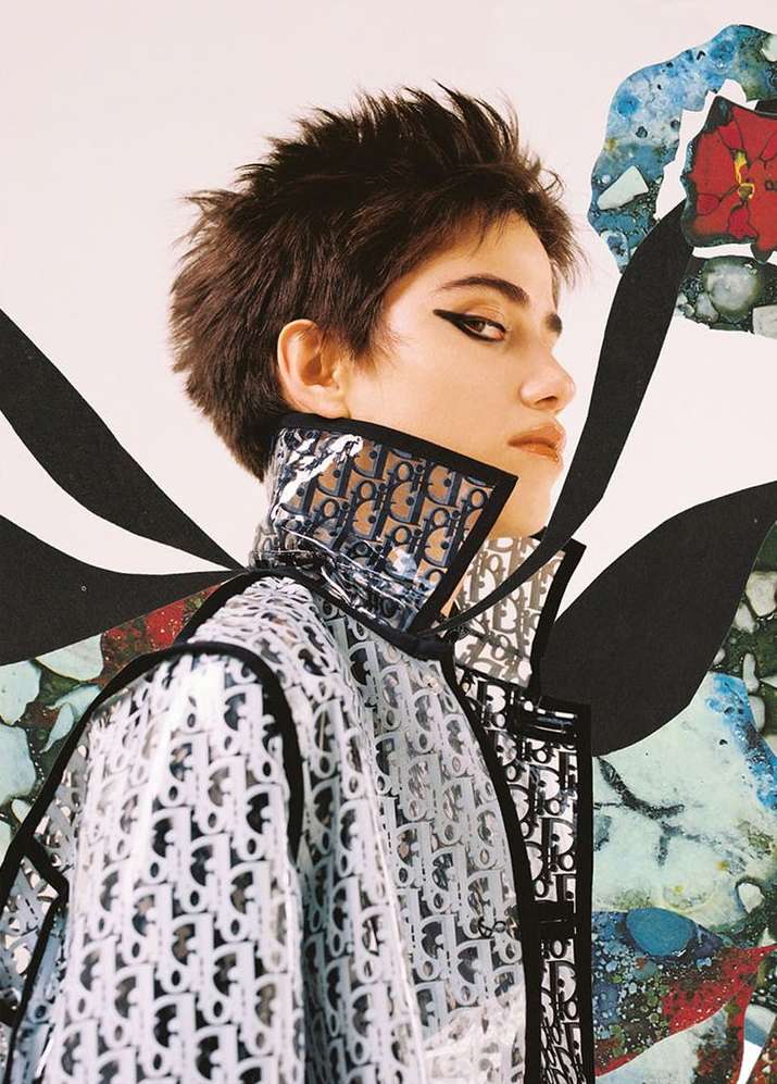 Woman with short brunette hair poses in translucent Dior-branded jacket against an abstract floral background in an indoor setting.