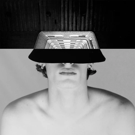 Black and white portrait of a shirtless man wearing a structural, geometric hat casting a shadow over his face, artistic composition.