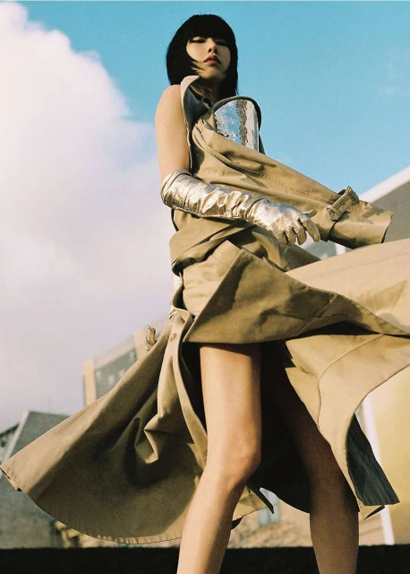 Woman in futuristic fashion posing outdoors with silver metallic gloves and top, and beige trench coat with blue sky in background.