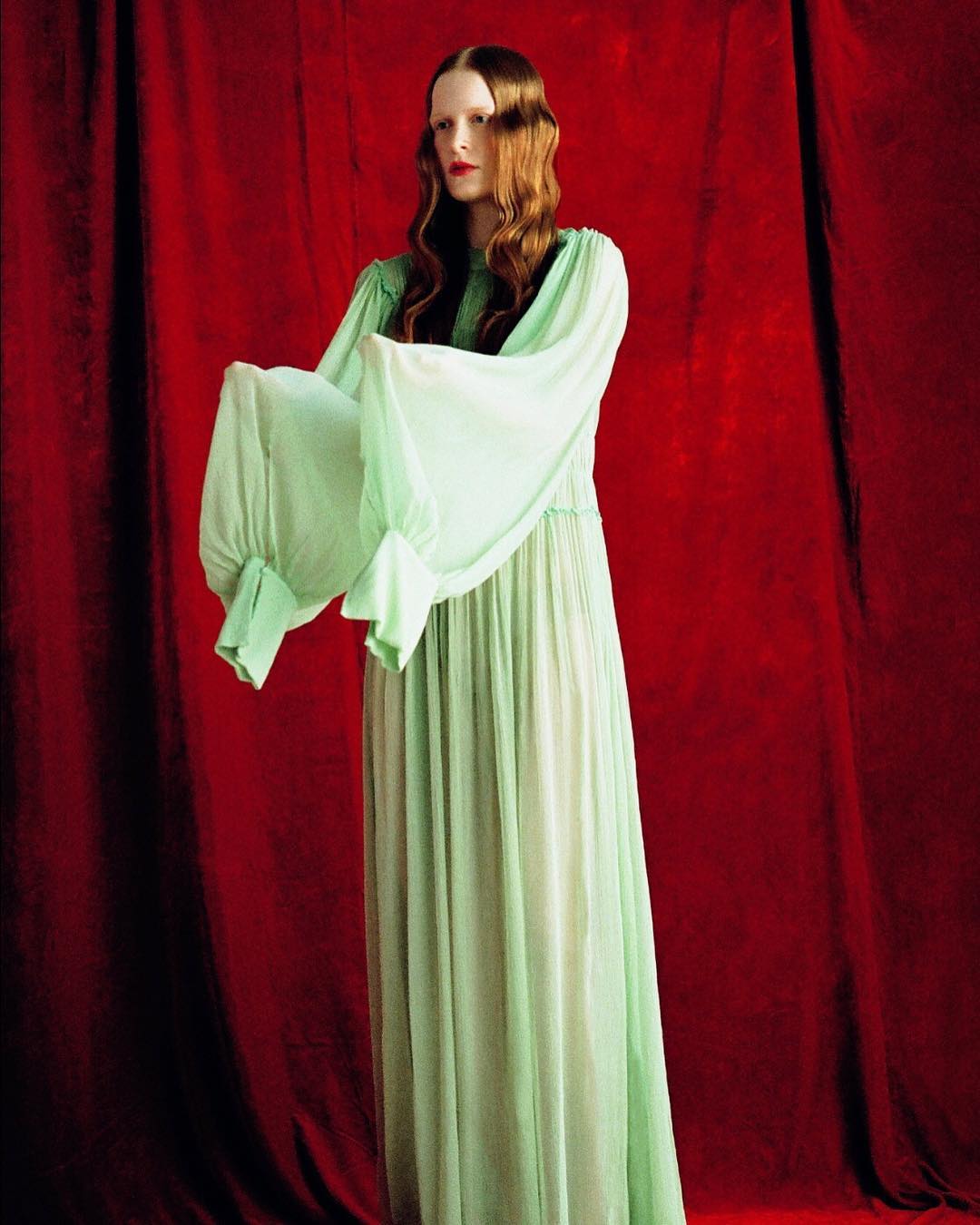 A fair-skinned woman with auburn hair in a light green gown poses against a red curtain backdrop in an indoor setting.