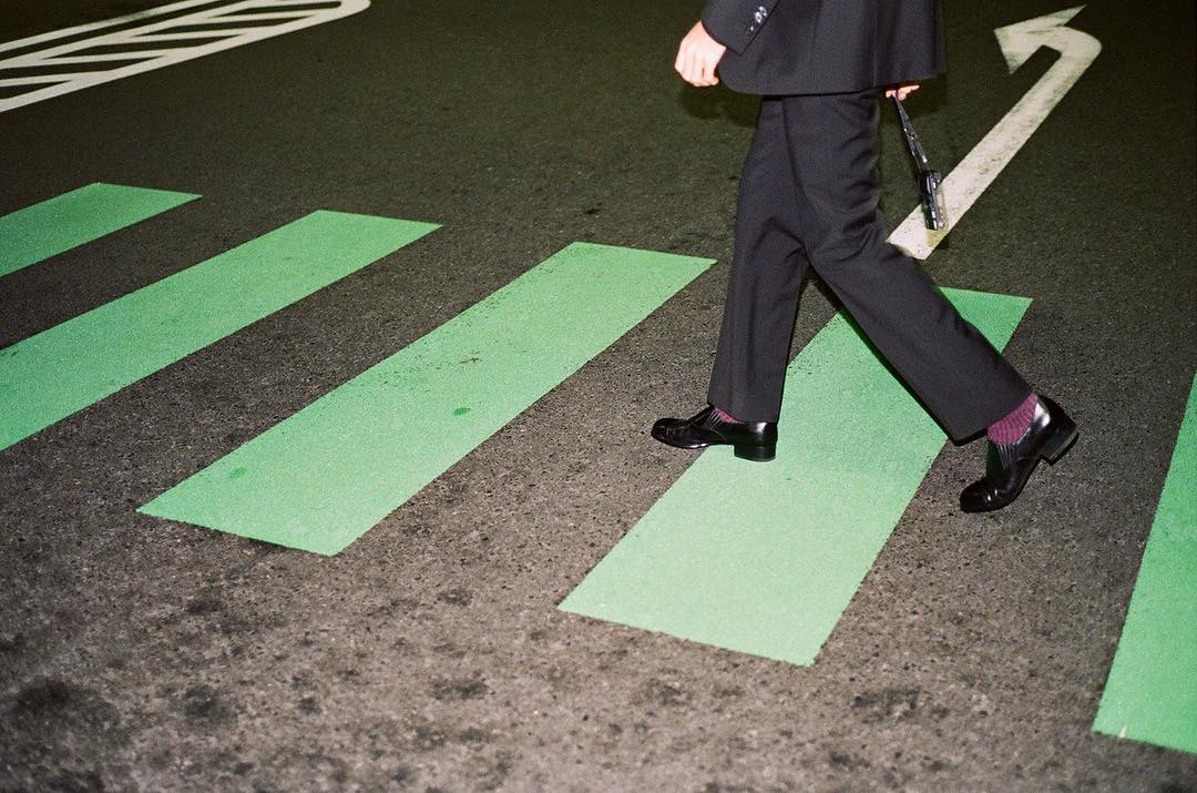 A person in a suit walks across a green and gray crosswalk at night, with a white arrow painted on the street.
