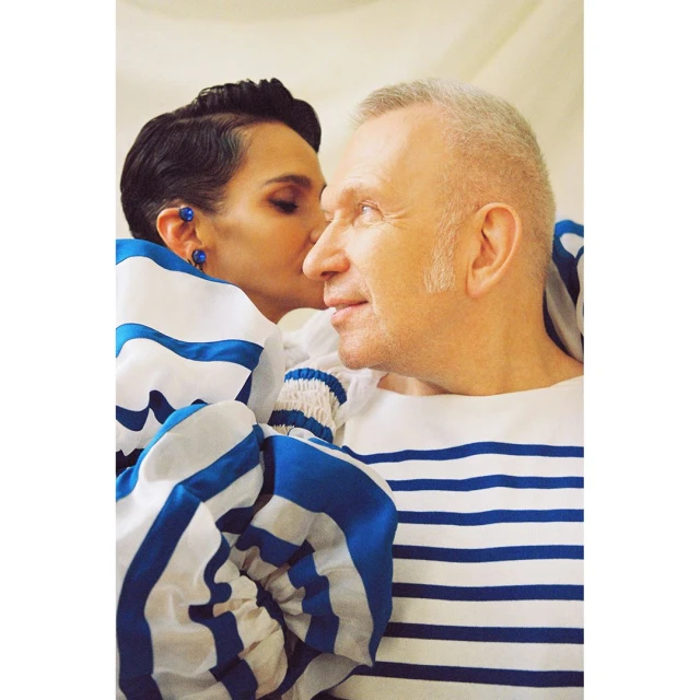 Close-up shot of a woman with short dark hair whispering in the ear of a smiling older man with salt-and-pepper hair, both wearing striped tops.