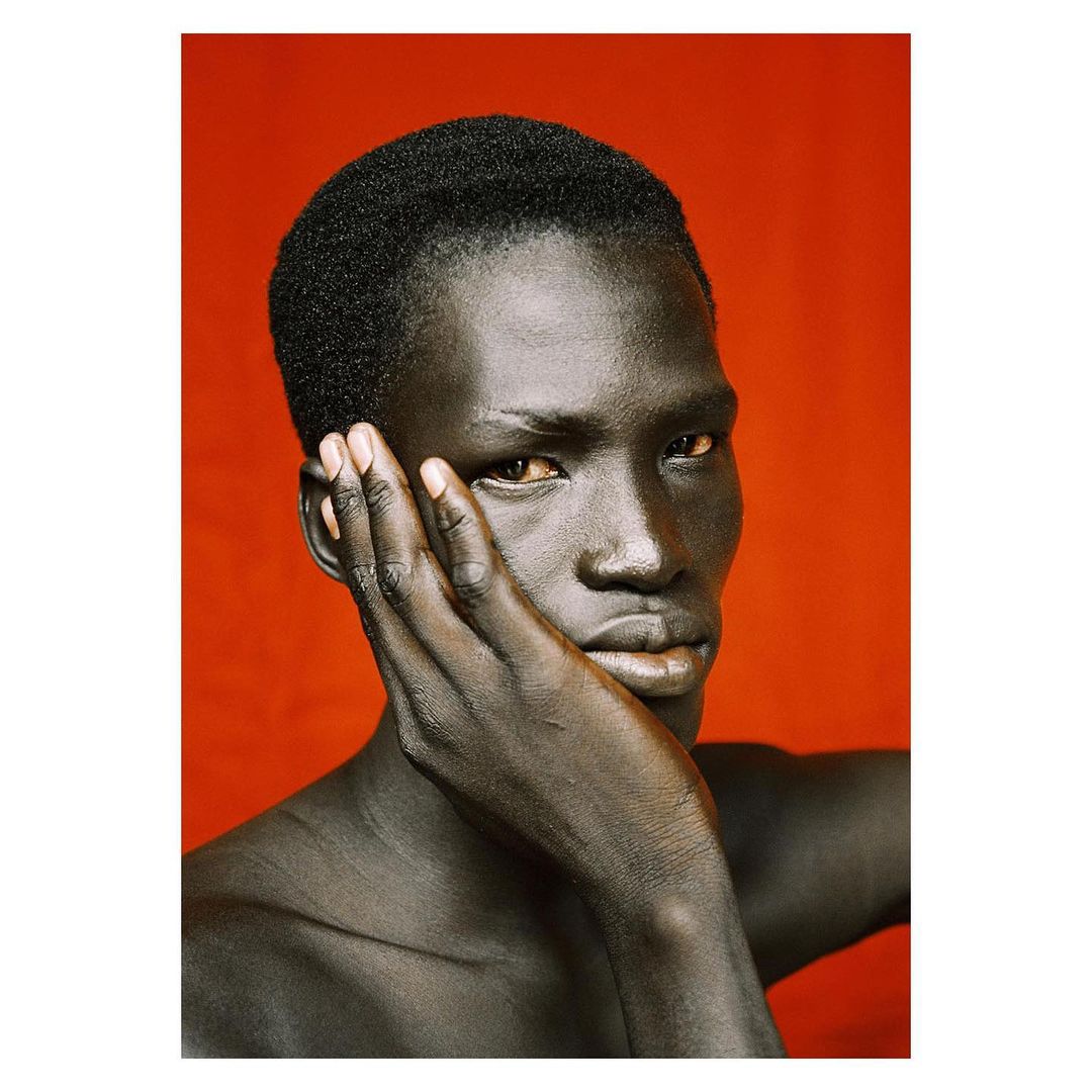Close-up portrait of a young Black man holding his hand to his cheek against a vibrant red backdrop.