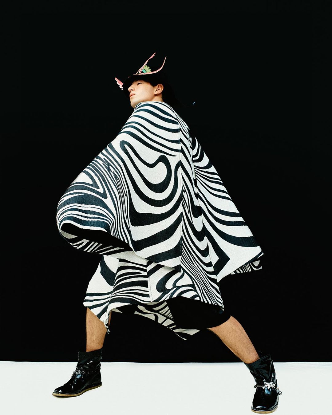An adult Asian male wearing a striped poncho and hat poses in front of a black backdrop with a white floor. Fashion editorial.