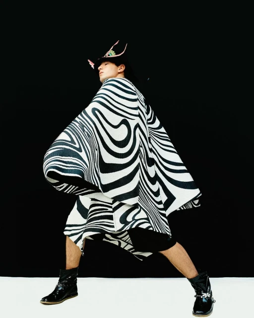 An adult Asian male wearing a striped poncho and hat poses in front of a black backdrop with a white floor. Fashion editorial.