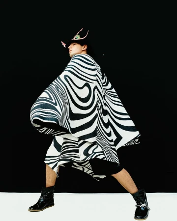 An adult Asian male wearing a striped poncho and hat poses in front of a black backdrop with a white floor. Fashion editorial.