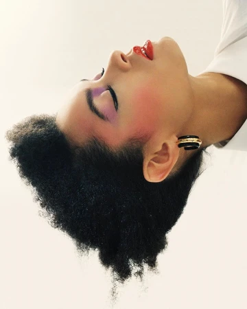 Profile portrait of a model with purple eyeshadow and red lips wearing black hoop earrings against a white background.