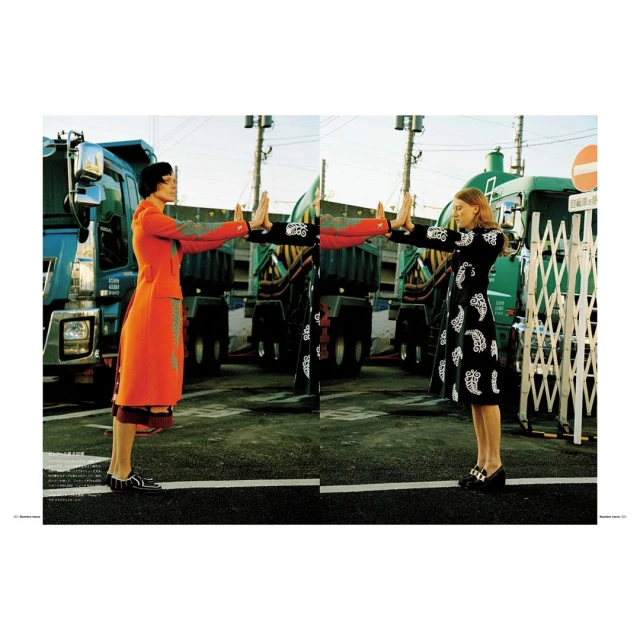 Two women in colorful dresses stand facing each other in front of large trucks on a city street in a fashion photography composition.