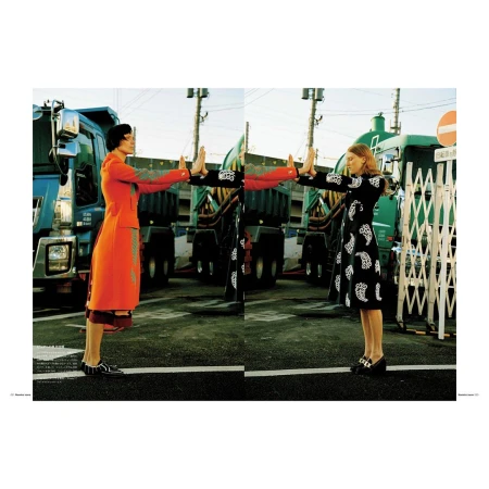 Two women in colorful dresses stand facing each other in front of large trucks on a city street in a fashion photography composition.