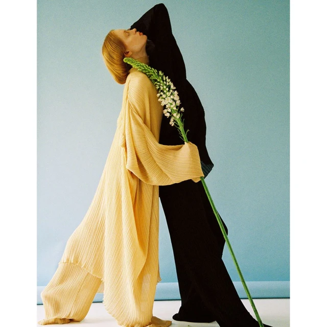 Two models in flowing garments pose with a flower stem against a pale blue backdrop in a studio fashion shot with neutral color grading.