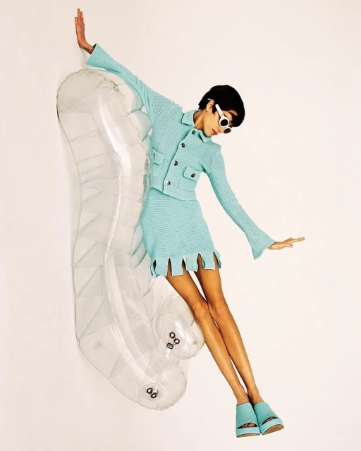 A fashion model with sunglasses, wearing a matching aqua blue skirt suit and platform shoes, poses with a clear inflatable chaise lounge.