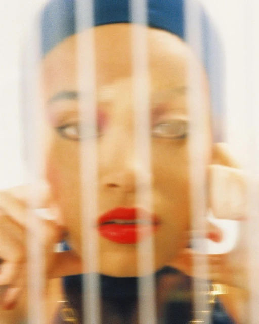 Woman with bright red lipstick and pink eyeshadow poses with hands framing face, distorted by blurry glass in a beauty fashion shot.