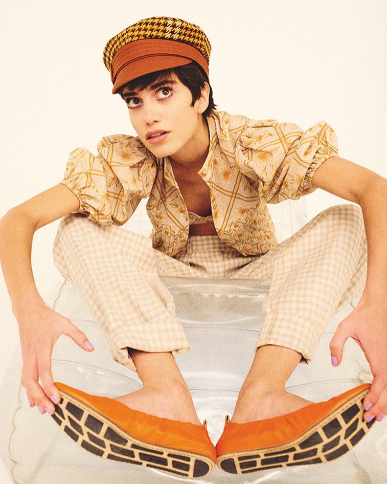 A young adult woman sits on a transparent surface, wearing a houndstooth newsboy cap, patterned top and pants, and orange flats with black soles.