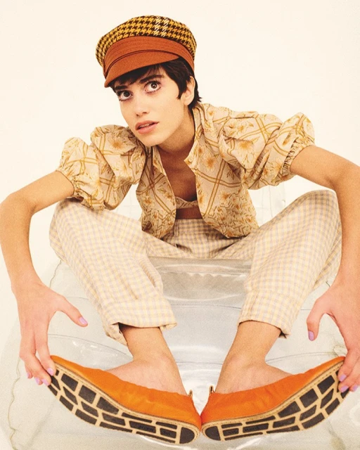 A young adult woman sits on a transparent surface, wearing a houndstooth newsboy cap, patterned top and pants, and orange flats with black soles.