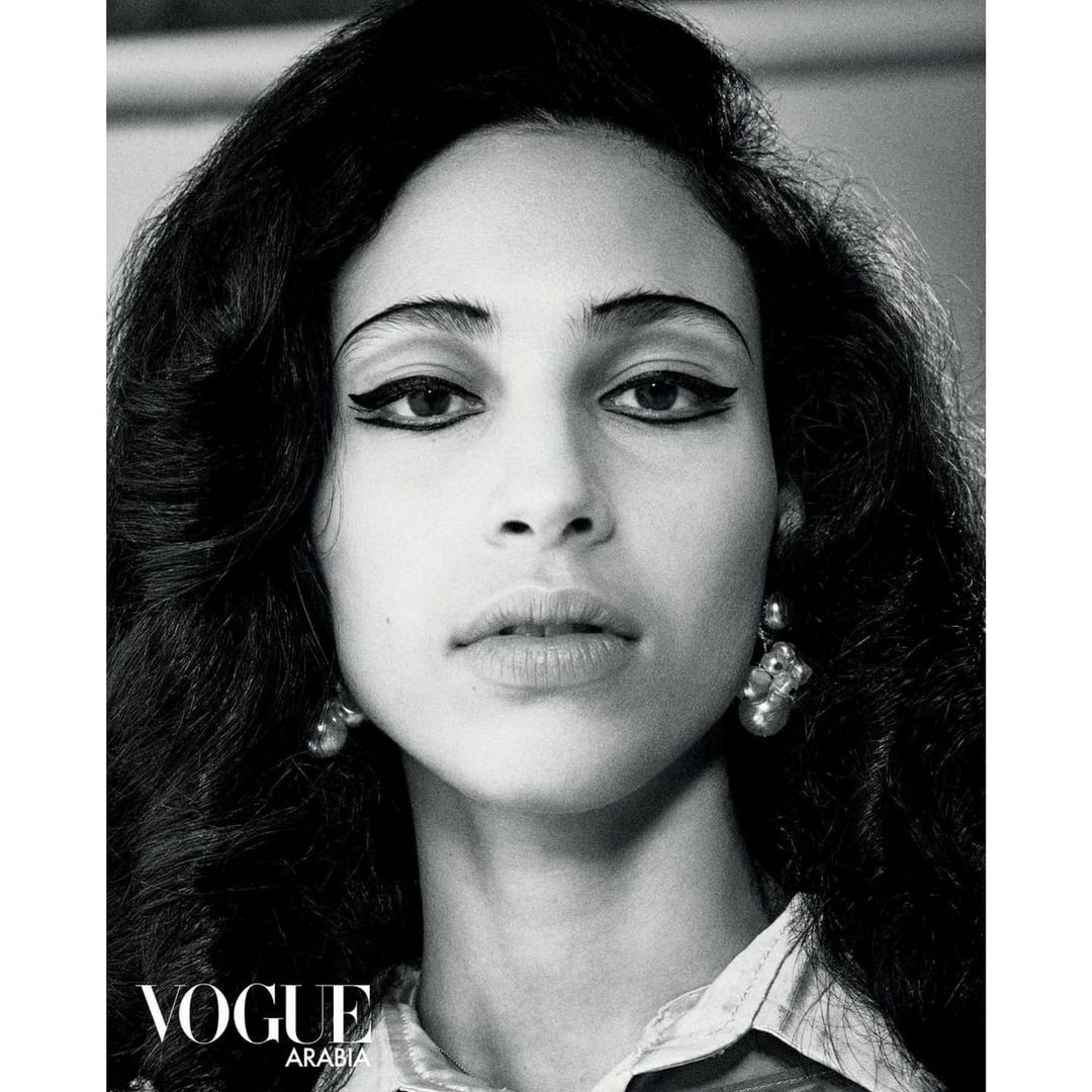 Black and white portrait of a woman with dark, voluminous hair and winged eyeliner, wearing drop earrings and a patterned top.