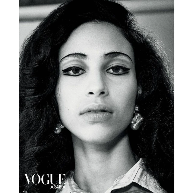 Black and white portrait of a woman with dark, voluminous hair and winged eyeliner, wearing drop earrings and a patterned top.