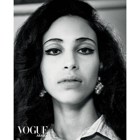 Black and white portrait of a woman with dark, voluminous hair and winged eyeliner, wearing drop earrings and a patterned top.