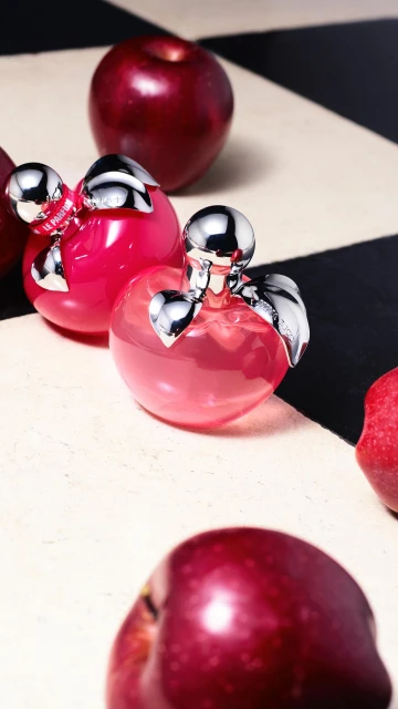 Product shot of two perfume bottles resembling apples, featuring reflective silver caps and pink liquids, surrounded by red apples.