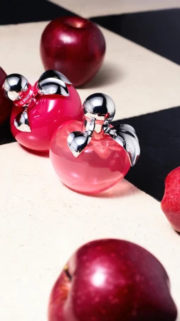 Product shot of two perfume bottles resembling apples, featuring reflective silver caps and pink liquids, surrounded by red apples.