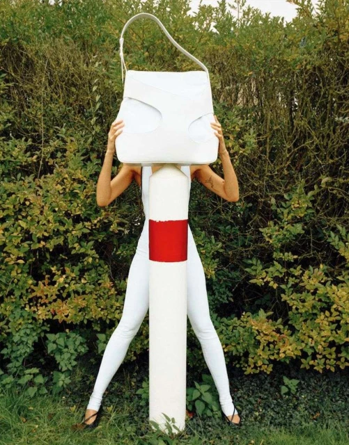 Person in white stands behind a white and red pole holding a large white handbag against a green hedge backdrop.