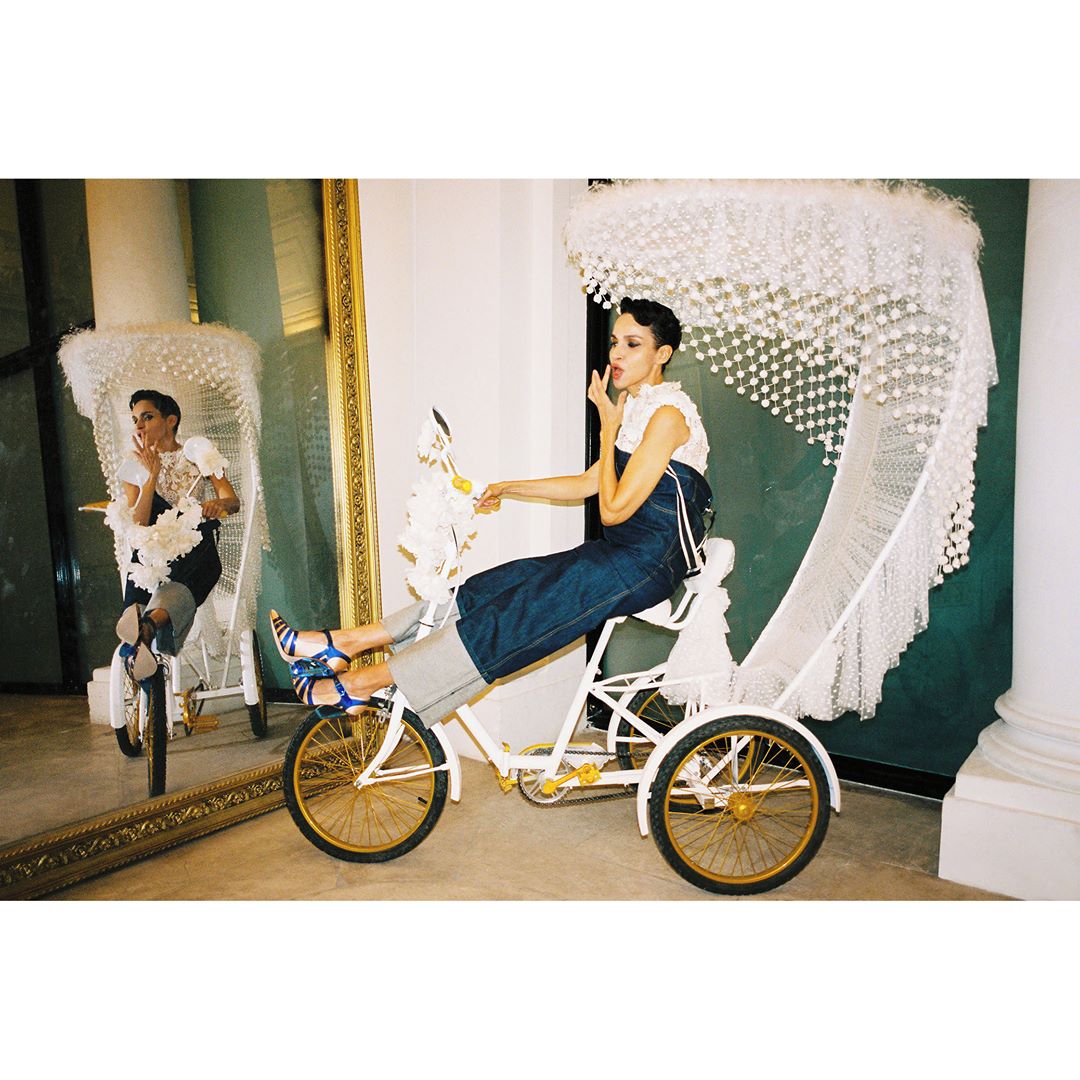 Woman in white top and denim pants sits sideways on a vintage tricycle with an ornate canopy, reflected in a gold-framed mirror.