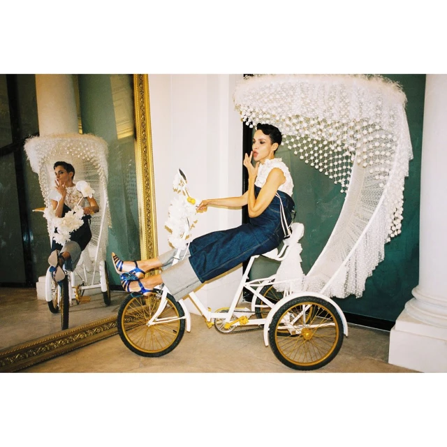 Woman in white top and denim pants sits sideways on a vintage tricycle with an ornate canopy, reflected in a gold-framed mirror.
