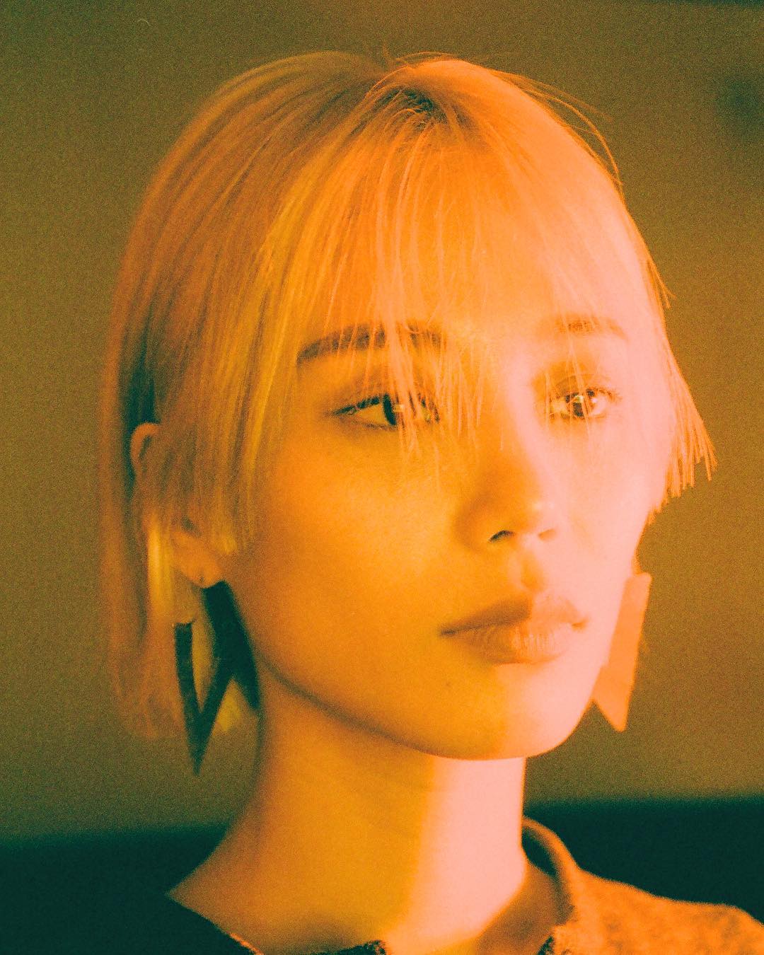Close-up portrait of a young adult female with short blonde hair, angular earrings, looking forward with soft lighting and warm tones.
