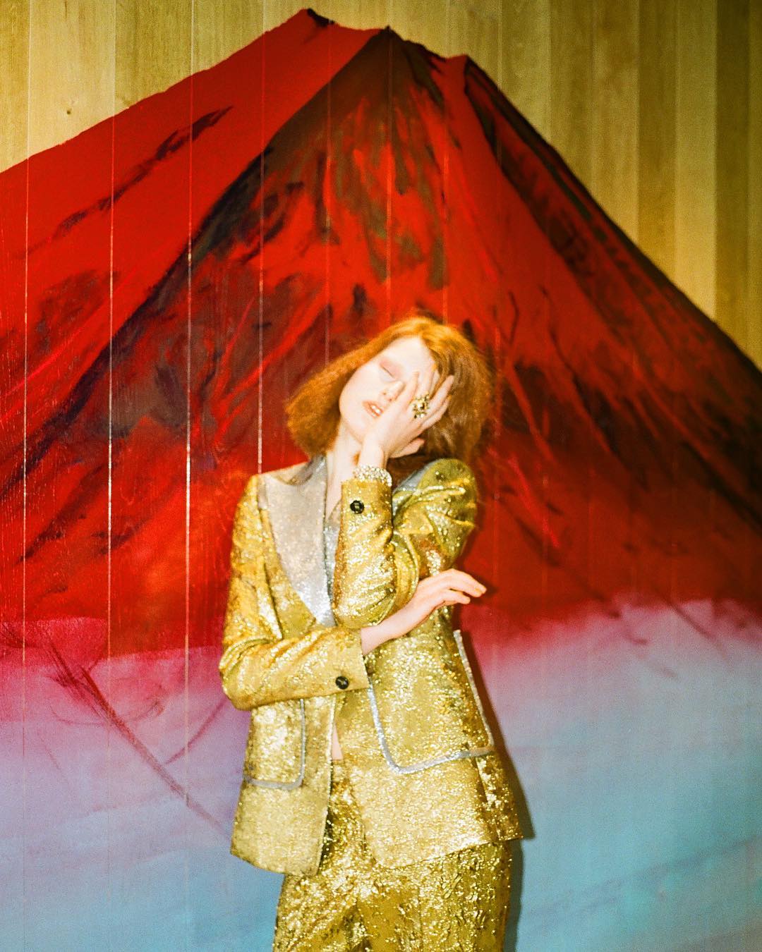 An adult female in a gold sequin suit stands in front of a backdrop with a red and black mountain painting, creating a high-fashion portrait.