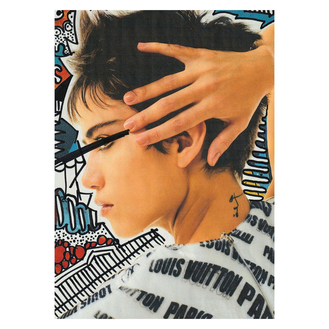 Close-up of a woman with short hair applying mascara against a colorful graphic art background with fashion brand text.