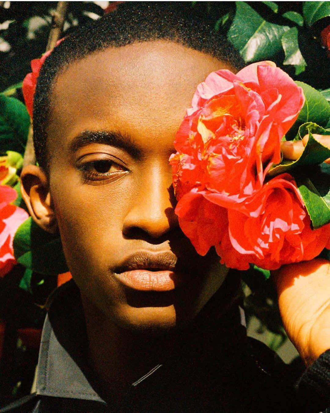 Portrait of a young Black man with a striking gaze, framed by vibrant red camellia flowers against a backdrop of lush green leaves.