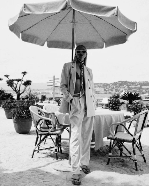 Woman in a blazer and trousers standing under a beach umbrella near outdoor cafe seating with a cityscape in the background in black and white.