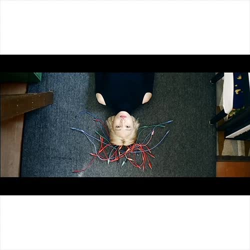 Woman with blonde hair lies on her back on a grey carpet with colorful wires around her head. The angle is shot from above.