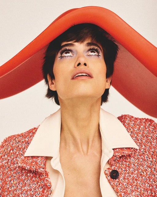 Portrait of a young woman with short dark hair, fair skin, and heavy makeup, looking up while wearing a red jacket and red head covering.