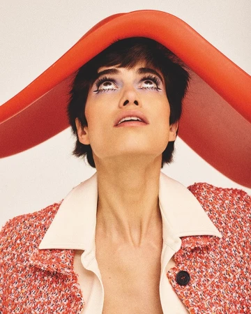 Portrait of a young woman with short dark hair, fair skin, and heavy makeup, looking up while wearing a red jacket and red head covering.