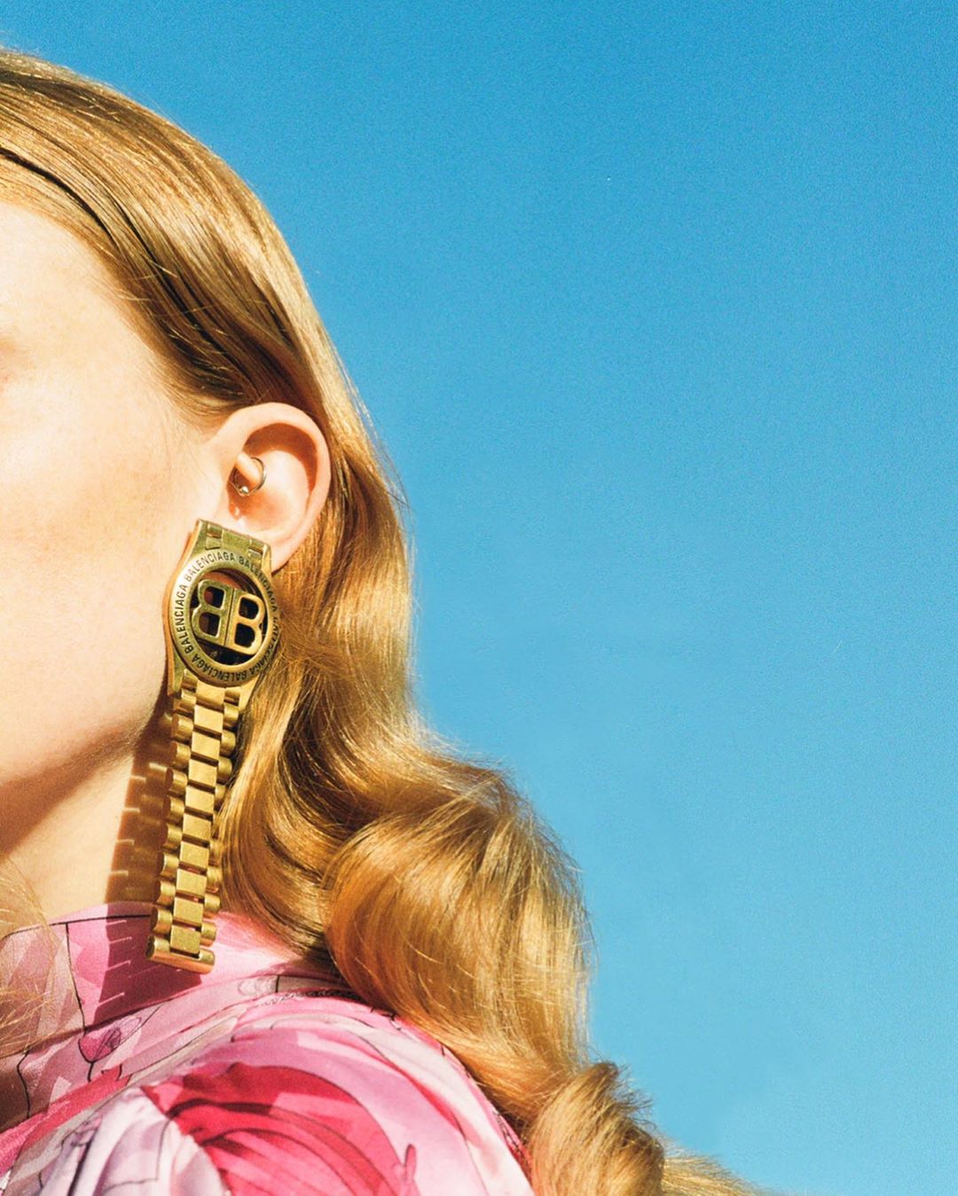 Close-up of a blonde woman with large gold Balenciaga BB earrings resembling wristwatches against a bright blue sky backdrop.