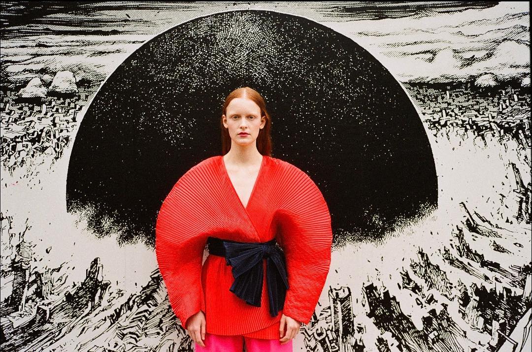 Fashion shot of a red-haired model in a pleated red outfit with a black obi belt, posing in front of a black and white illustrated backdrop.