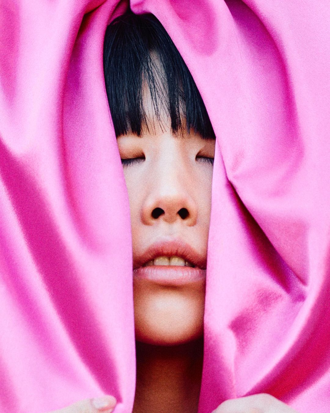 Close-up portrait of a young adult with closed eyes, framed by draped pink fabric, creating a soft and intimate aesthetic.