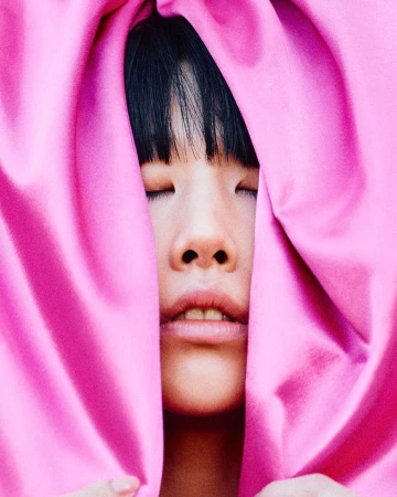 Close-up portrait of a young adult with closed eyes, framed by draped pink fabric, creating a soft and intimate aesthetic.
