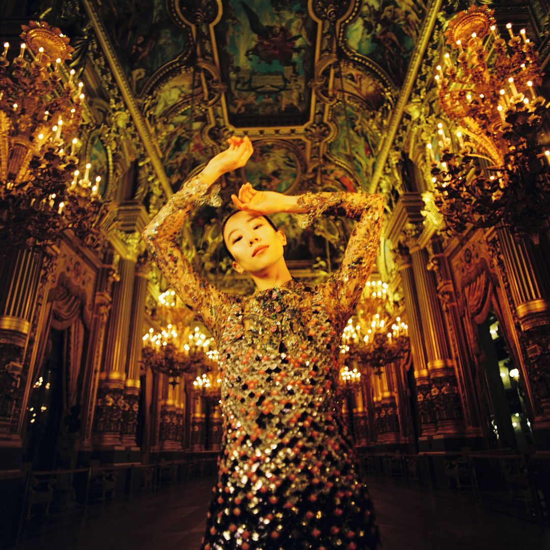 Elegant Asian woman in shimmering gold dress poses in an ornate ballroom with chandeliers and ceiling art. Fashion, glamour, style.